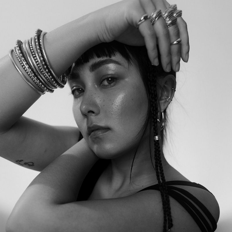 Woman with silver bangles and rings poses with arms crossed over her face, showing multiple ear piercings and a braided hair strand.