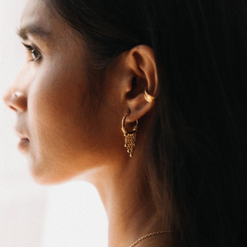 Close-up side profile of a woman wearing intricate gold hoop earrings with chain details and a smaller gold ear cuff, softly lit by natural light.