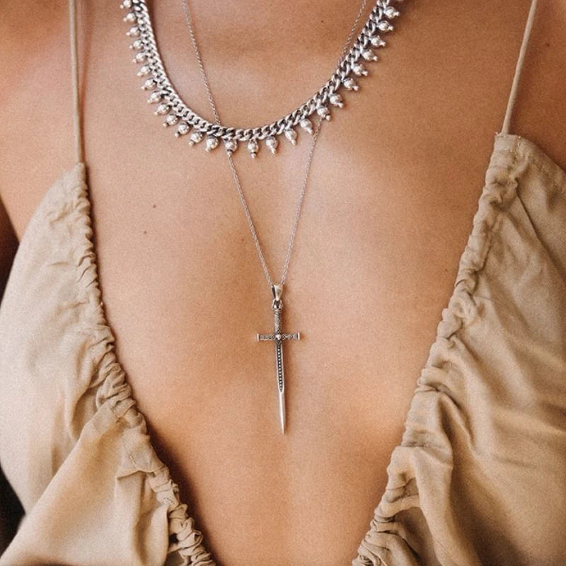 Silver sword pendant necklace on a long fine chain, layered beneath a beaded curb chain choker, the blade resting flat at the center of the chest