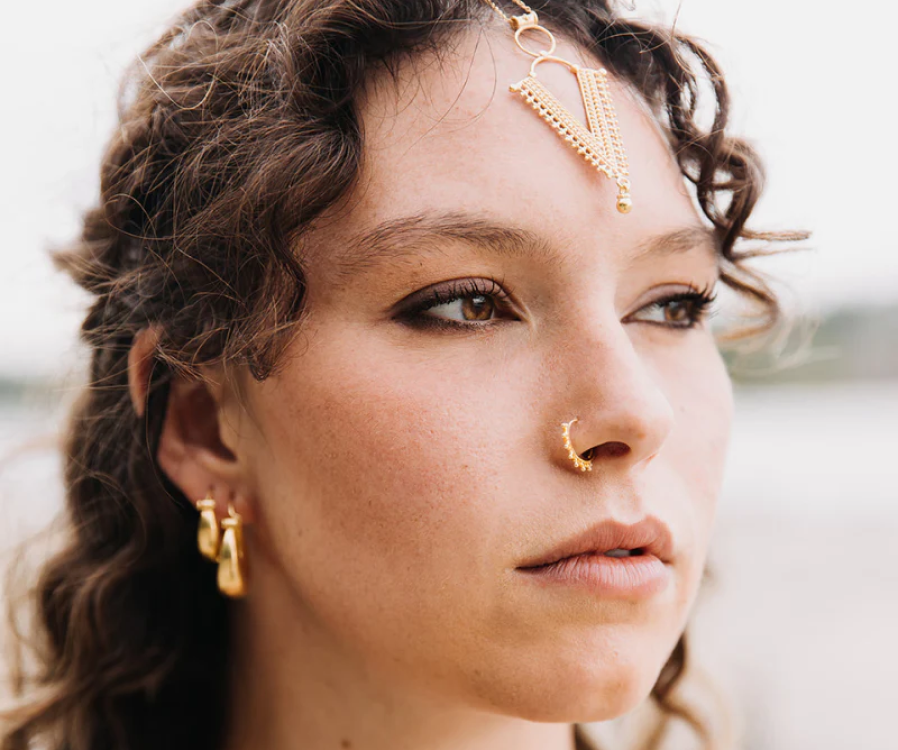 Gold beaded septum ring worn with a fine chained headpiece and small gold hoop earrings, jewelry framing the center of the face